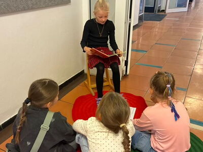 Eine Schülerin sitzt auf einem Stuhl und liest anderen Schülerinnen daraus vor